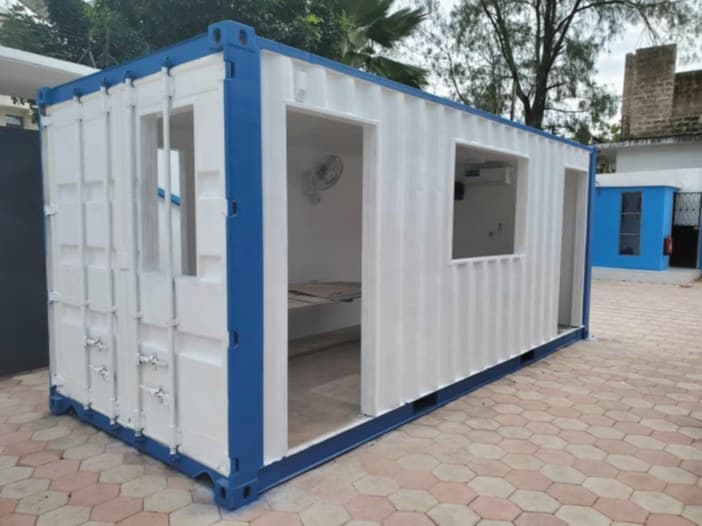 A white shipping container converted into an office, with blue frame, windows and an open door, on a paved yard.