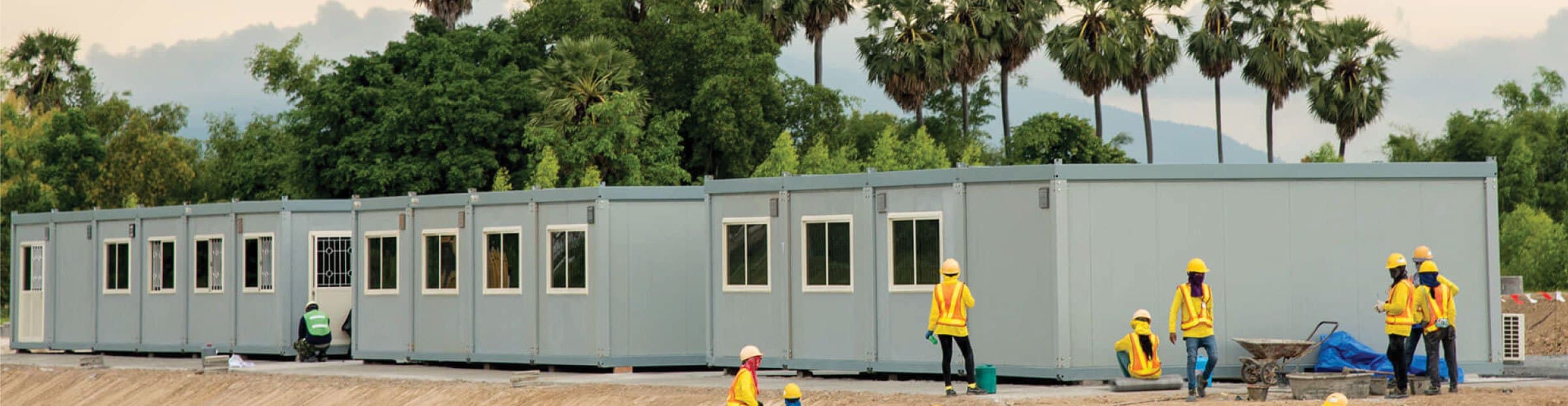 A row of grey prefabricated site buildings being installed by a crew in hard hats and high-visibility vests.