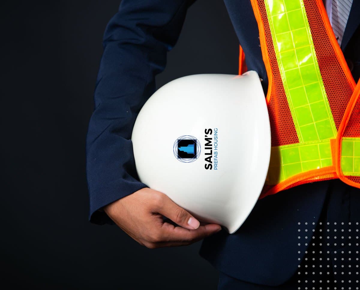 A site lead in a suit and high-visibility vest holding a white hard hat with the Salim's Prefab Housing logo.